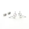 Ett par "Örhänge med stift - Serotonin molekyl" silverörhängen, utformade som en serotoninmolekyl, med två fjärilsryggar bredvid på en vit bakgrund.