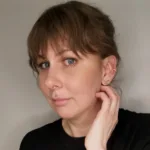 En person med ljus hy och blå ögon tittar in i kameran och tar sig för ansiktet med ena handen. Hen har brunt hår med lugg, är lätt sminkad, har en läppiercing och två örhängen. Bakgrunden är enkel och neutral.