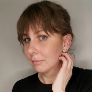 En person med ljus hy och blå ögon tittar in i kameran och tar sig för ansiktet med ena handen. Hen har brunt hår med lugg, är lätt sminkad, har en läppiercing och två örhängen. Bakgrunden är enkel och neutral.