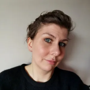 En person med kort brunt hår och blå ögon, klädd i en svart tröja, tittar in i kameran mot en vanlig ljus bakgrund.