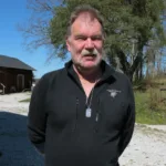En medelålders man med mustasch står utomhus iklädd en svart jacka med dragkedja och Hängsmycke Bricka AMS 800 i sterlingsilverhalsband. Bakom honom syns träd, en röd byggnad och en grusväg en solig dag.