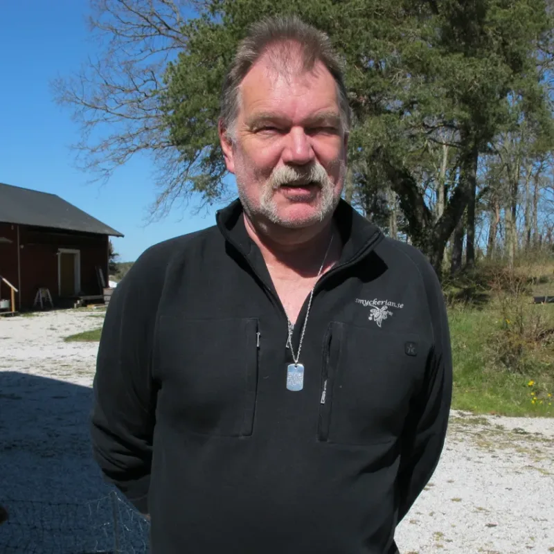 En medelålders man med mustasch står utomhus iklädd en svart jacka med dragkedja och Hängsmycke Bricka AMS 800 i sterlingsilverhalsband. Bakom honom syns träd, en röd byggnad och en grusväg en solig dag.