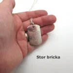 En hand håller i Hängsmycke Bricka AMS 800 i sterlingsilver, ett halsband av silver med graverad text, mot en vit bakgrund. Under handen finns texten "Stor bricka" (på svenska).