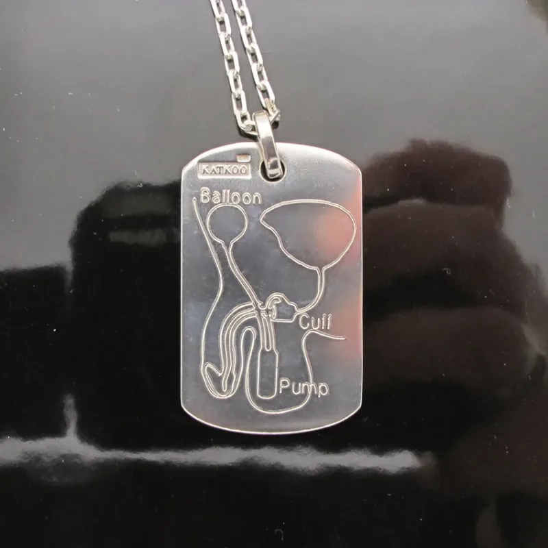Hängsmycke Bricka AMS 800 i sterlingsilver är ett silverhalsband med ett graverat diagram över ett penisimplantat märkt Ballong, Manschett och Pump, mot en blank svart bakgrund.