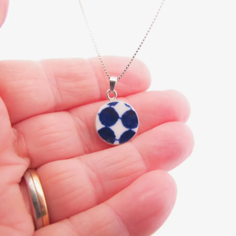 En hand håller i ett Porslinssmycke Adam 22x15mm halsband med ett runt blåvitt prickigt hänge. Bakgrunden är vit och personen bär en guld- och silverring.