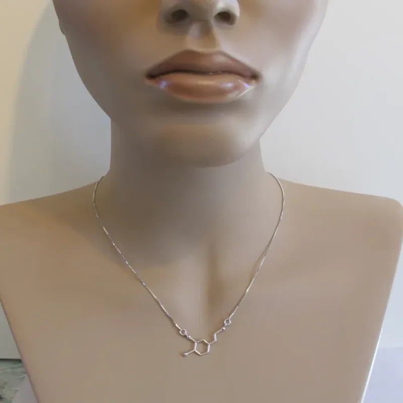 En skyltdocka visar Silverhalsbandets molekylen för Dopamin - ett delikat kedjehalsband med en dopaminmolekyl som hänge - mot en slät bakgrund.