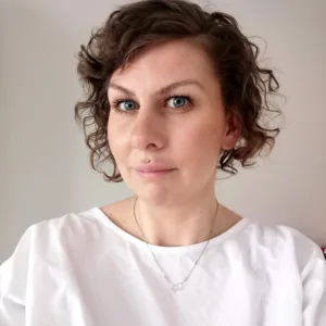 En kvinna med kort, lockigt brunt hår och blå ögon bär en vit topp och Silverhalsbandets halsband molekylen för Serotonin och tittar in i kameran med ett neutralt uttryck mot en vanlig ljus bakgrund.