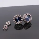 Örhängena Örhänge med stift Krona - Adam, med blå- och vitmönstrade cirkelformade mittringar i utsmyckad silverfärgad metall, visas på en reflekterande svart yta med fjärilsryggar bredvid.