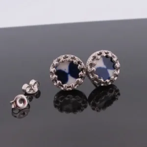 Örhängena Örhänge med stift Krona - Adam, med blå- och vitmönstrade cirkelformade mittringar i utsmyckad silverfärgad metall, visas på en reflekterande svart yta med fjärilsryggar bredvid.