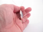 Närbild av en hand som håller i "Halsband med handslipad Kalcedon 38x25mm", med ett silverfärgat halsbandshänge med en grå kalcedonsten, från sidan mot en vit bakgrund.