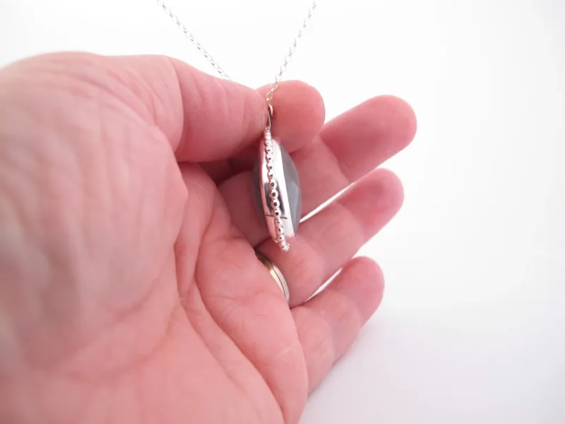 Närbild av en hand som håller i "Halsband med handslipad Kalcedon 38x25mm", med ett silverfärgat halsbandshänge med en grå kalcedonsten, från sidan mot en vit bakgrund.