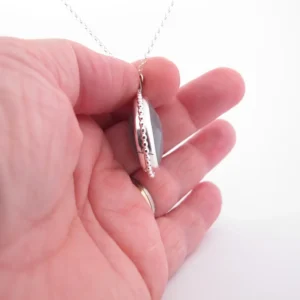 Närbild av en hand som håller i "Halsband med handslipad Kalcedon 38x25mm", med ett silverfärgat halsbandshänge med en grå kalcedonsten, från sidan mot en vit bakgrund.