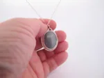 En hand håller i "Halsband med handslipad Kalcedon 38x25mm", med en oval grå kalcedon som hänge i en dekorativ silverram, mot en vit bakgrund.