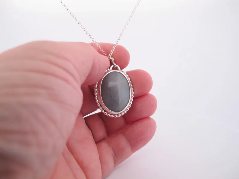 En hand håller i "Halsband med handslipad Kalcedon 38x25mm", med en oval grå kalcedon som hänge i en dekorativ silverram, mot en vit bakgrund.