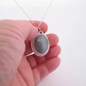 En hand håller i "Halsband med handslipad Kalcedon 38x25mm", med en oval grå kalcedon som hänge i en dekorativ silverram, mot en vit bakgrund.