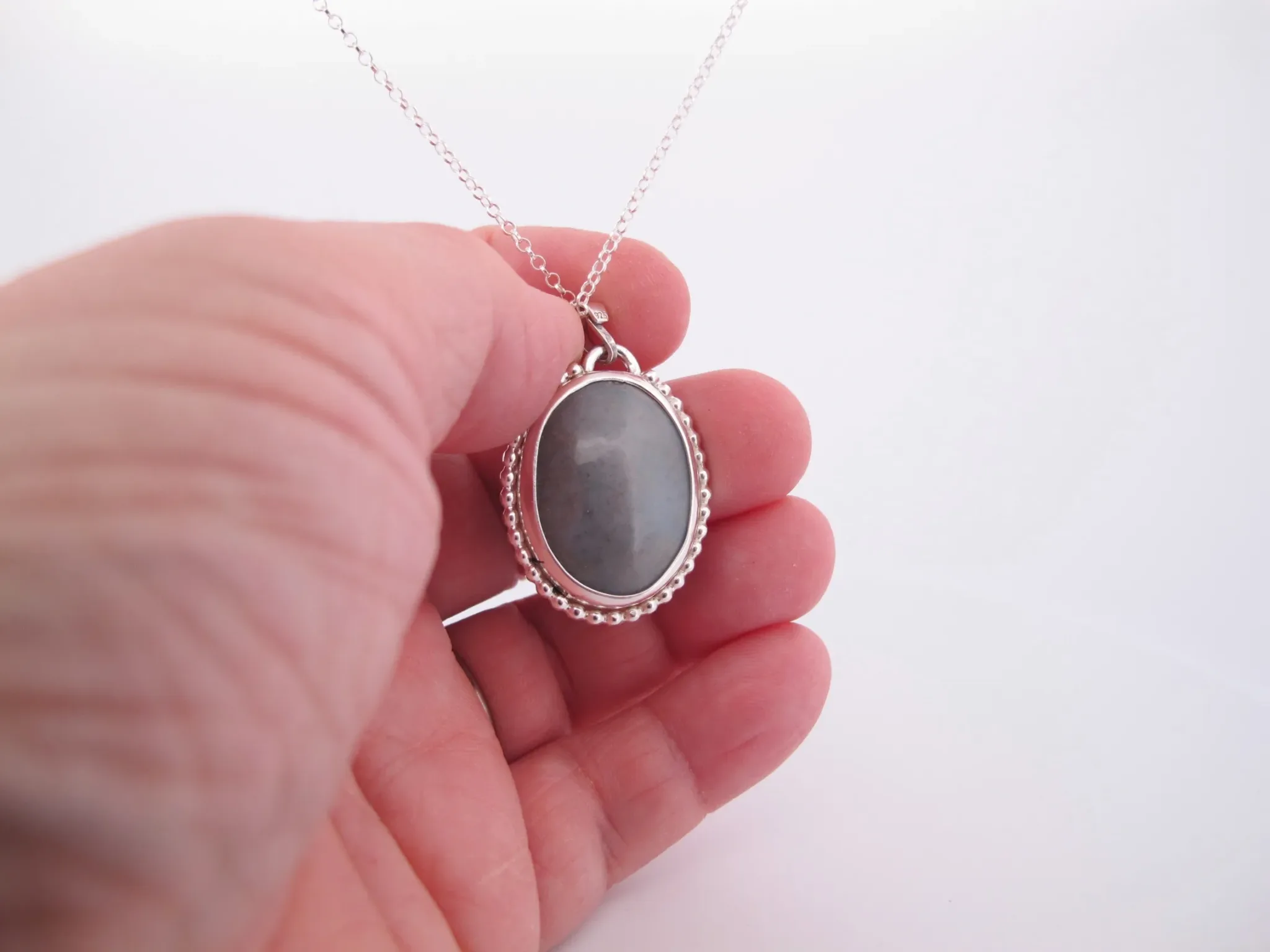 En hand håller i "Halsband med handslipad Kalcedon 38x25mm", med en oval grå kalcedon som hänge i en dekorativ silverram, mot en vit bakgrund.