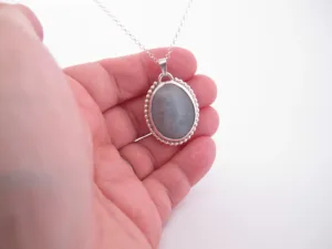 En hand håller i "Halsband med handslipad Kalcedon 38x25mm", ett silverhalsband med en oval grå kalcedon som hänge och en pärlbeströdd ram, mot en vit bakgrund.