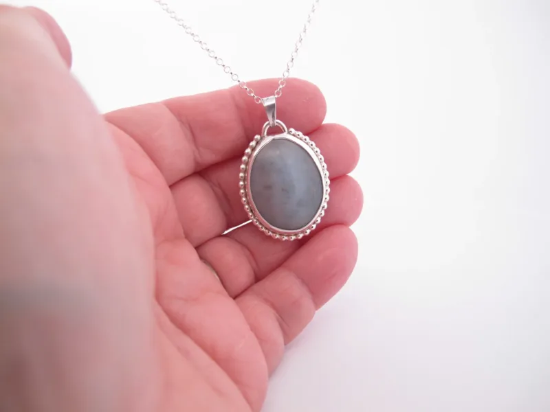 En hand håller i "Halsband med handslipad Kalcedon 38x25mm", ett silverhalsband med en oval grå kalcedon som hänge och en pärlbeströdd ram, mot en vit bakgrund.