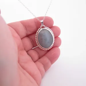 En hand håller i "Halsband med handslipad Kalcedon 38x25mm", ett silverhalsband med en oval grå kalcedon som hänge och en pärlbeströdd ram, mot en vit bakgrund.