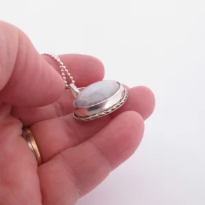 En hand håller i "Halsband med handslipad Kalcedon 32x23mm", ett silversmycke med en kupolformad, handslipad kalcedon i en vriden ram på en pärlkedja, mot en vit bakgrund.