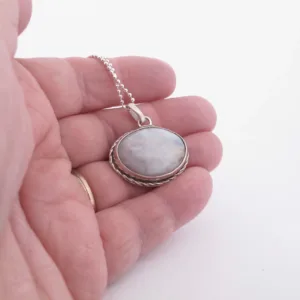 En hand håller i "Halsband med handslipad Kalcedon 32x23mm", ett silverhalsband med en rund, vitmarmorerad kalcedonsten i en dekorativ infattning, hängande i en tunn kedja mot en ljus bakgrund.