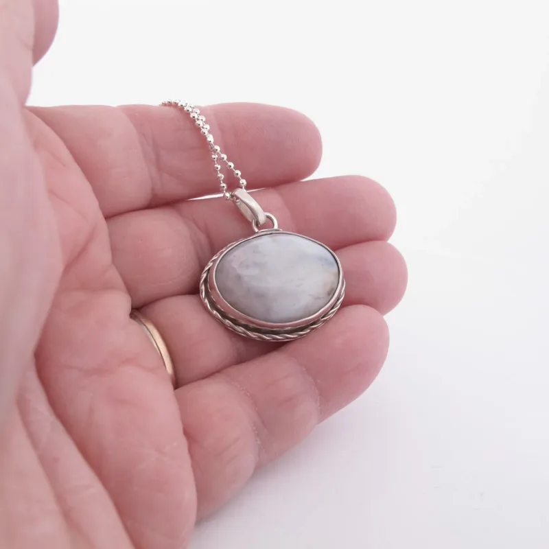 En hand håller i "Halsband med handslipad Kalcedon 32x23mm", ett silverhalsband med en rund, vitmarmorerad kalcedonsten i en dekorativ infattning, hängande i en tunn kedja mot en ljus bakgrund.