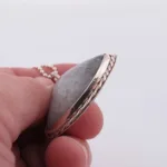 Närbild av en hand som håller i "Halsband med handslipad Kalcedon 45x40mm", ett grått ovalt stenhänge med silverkant och kedja, mot en vit bakgrund.