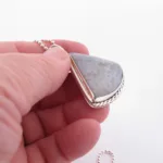 En hand håller i ett "Halsband med handslipad Kalcedon 45x40mm" - ett silverhänge med en triangulär grå kalcedonsten och snodd kant, på en pärlkedja, mot en vit bakgrund.