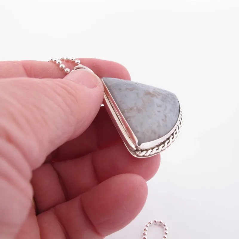 En hand håller i ett "Halsband med handslipad Kalcedon 45x40mm" - ett silverhänge med en triangulär grå kalcedonsten och snodd kant, på en pärlkedja, mot en vit bakgrund.