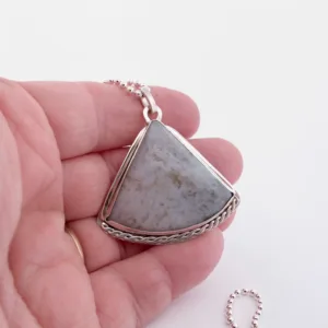 En hand håller i "Halsband med handslipad Kalcedon 45x40mm", ett triangulärt gråstenshänge med en silverfärgad tvinnad kant på ett kulkedjehalsband, mot en vit bakgrund.