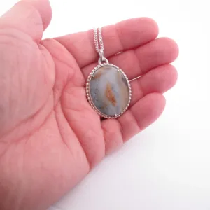 En hand håller i "Halsband med handslipad Kalcedon 45x30mm", med en oval, handpolerad kalcedonsten i en silverinfattning på en silverkedja, mot en vit bakgrund.