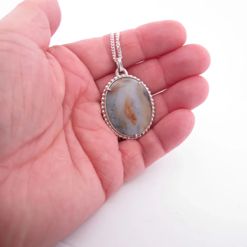 En hand håller i "Halsband med handslipad Kalcedon 45x30mm", med en oval, handpolerad kalcedonsten i en silverinfattning på en silverkedja, mot en vit bakgrund.