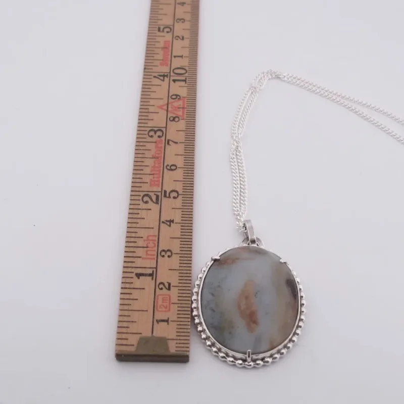 Ett silverhalsband med ett handskuret 45x30mm Chalcedon-hänge (marmorerat grått, vitt och brunt) ligger vid en trälinjal, som visar att hänget är ca 4 cm långt. Produkt: Halsband med handslipad Kalcedon 45x30mm.