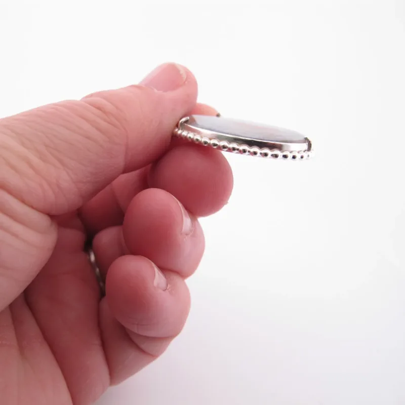 Närbild av en hand som håller i Halsband med handslipad Kalcedon 45x30mm - ett runt hänge med silverkant och pärlkant - sedd från sidan mot en vit bakgrund.