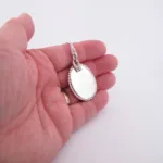 En hand håller i "Halsband med handslipad Kalcedon 45x30mm", ett ovalt silversmycke med pärlkant och matchande kedja, mot en vit bakgrund.