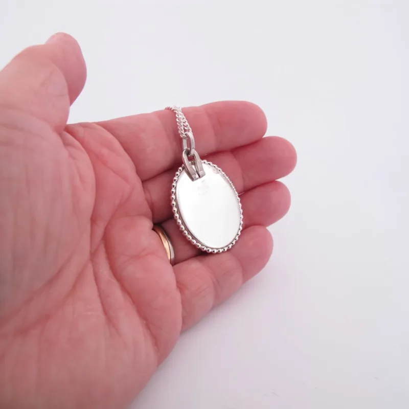 En hand håller i "Halsband med handslipad Kalcedon 45x30mm", ett ovalt silversmycke med pärlkant och matchande kedja, mot en vit bakgrund.
