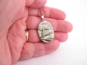 En hand håller i Porslinsmycke - Gröna Anna, ett ovalt silverhänge med grönt och vitt botaniskt mönster mot en vit bakgrund.