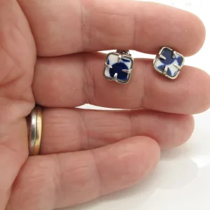Närbild av en hand som håller i två "Örhänge med stift - Adam"-örhängen: små kvadrater med blå kors, vita accenter och silverramar. Personen bär en vigselring i guld.