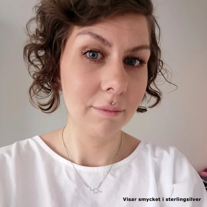 En person med kort, lockigt brunt hår och blå ögon bär en vit topp och Halsbandsmolekylen för Dopamin i sterling silver. De har en diskret makeup, en näspiercing, en slät bakgrund och text: "Visar smycket i sterlingsilver.