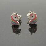 Ett par "Örhänge med stift - Enhörning" enhörningsformade stiftörhängen med vita ansikten, rosa manar och regnbågsdetaljer visas på en reflekterande svart yta.