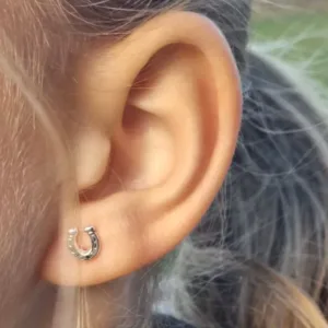 Närbild på en persons öra som bär Örhänge med stift - Enhörning, ett litet enhörningsörhänge. Håret är bakåtkammat och bakgrunden visar suddiga gröna och bruna toner.