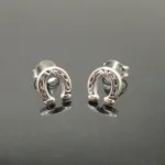 Ett par små enhörningsörhängen i silver, kallade "Örhänge med stift - Enhörning", visas på en reflekterande mörk yta.