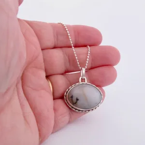En hand håller i "Halsband med handslipad Kalcedon", ett silverhalsband med ett ovalt hänge av vit kalcedon med subtila grå och svarta strimmor, mot en vit bakgrund.