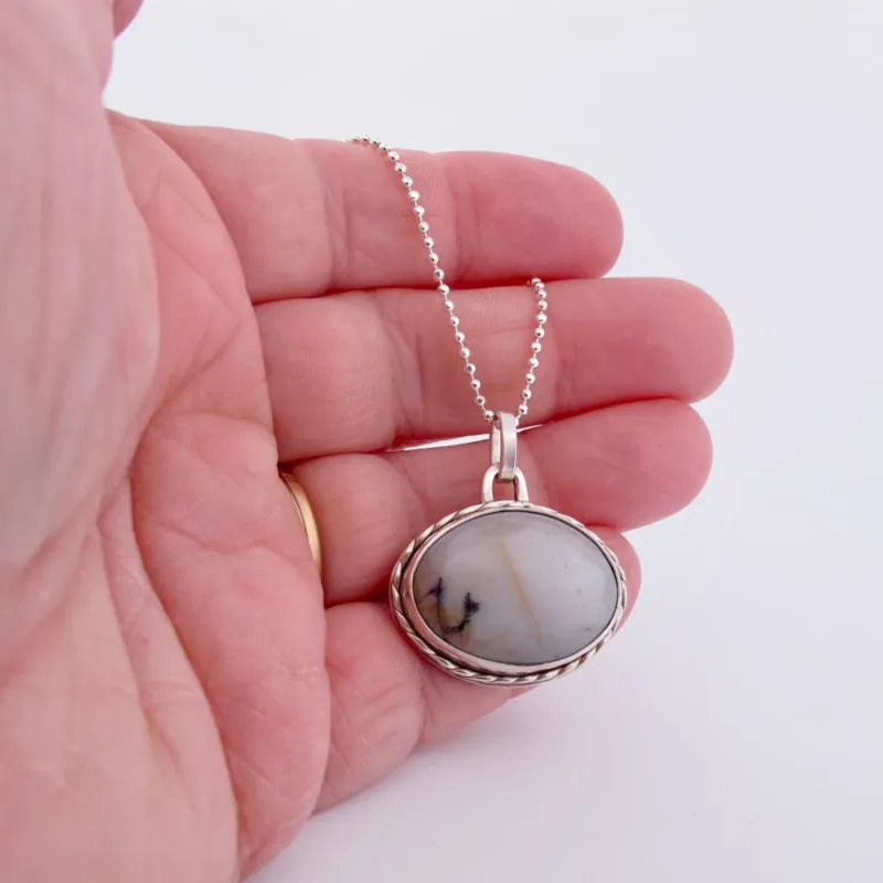 En hand håller i "Halsband med handslipad Kalcedon", ett silverhalsband med ett ovalt hänge av vit kalcedon med subtila grå och svarta strimmor, mot en vit bakgrund.