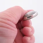 En hand håller i "Halsband med handslipad Kalcedon", med en rund, vit kalcedon med grå ådror infattad i en silverfärgad infattning med flätad kant, mot en vit bakgrund.
