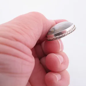 En hand håller i "Halsband med handslipad Kalcedon", med en rund, vit kalcedon med grå ådror infattad i en silverfärgad infattning med flätad kant, mot en vit bakgrund.