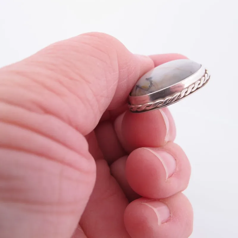 En hand håller i "Halsband med handslipad Kalcedon", med en rund, vit kalcedon med grå ådror infattad i en silverfärgad infattning med flätad kant, mot en vit bakgrund.