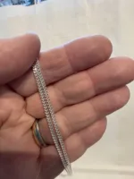 En hand håller i Silverkedja pansar 2.1mm halskedja (50-60cm) och visar dess geometriska, sammanlänkade silverkedjedesign. Personen bär också en guld- och silverring på sitt ringfinger, med en vanlig suddig bakgrund.