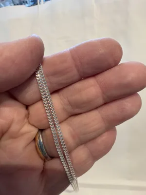 En hand håller i Silverkedja pansar 2.1mm halskedja (50-60cm) och visar dess geometriska, sammanlänkade silverkedjedesign. Personen bär också en guld- och silverring på sitt ringfinger, med en vanlig suddig bakgrund.