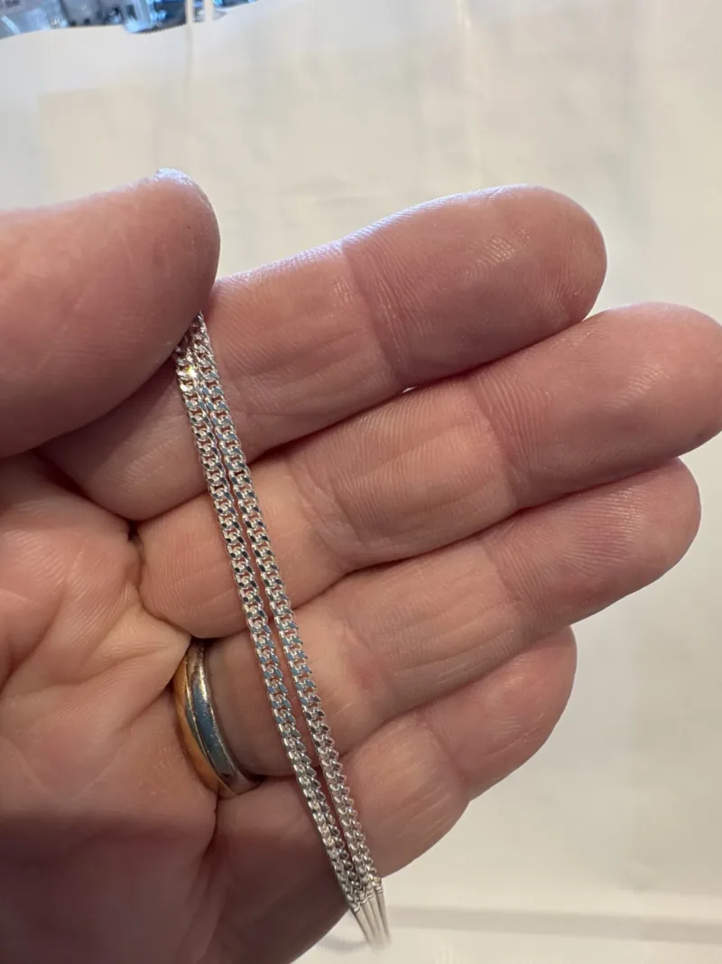 En hand håller i Silverkedja pansar 2.1mm halskedja (50-60cm) och visar dess geometriska, sammanlänkade silverkedjedesign. Personen bär också en guld- och silverring på sitt ringfinger, med en vanlig suddig bakgrund.